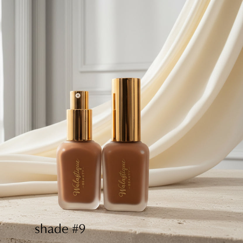 Two brown liquid foundation bottles by Walastique Beauty with gold caps on a textured surface.